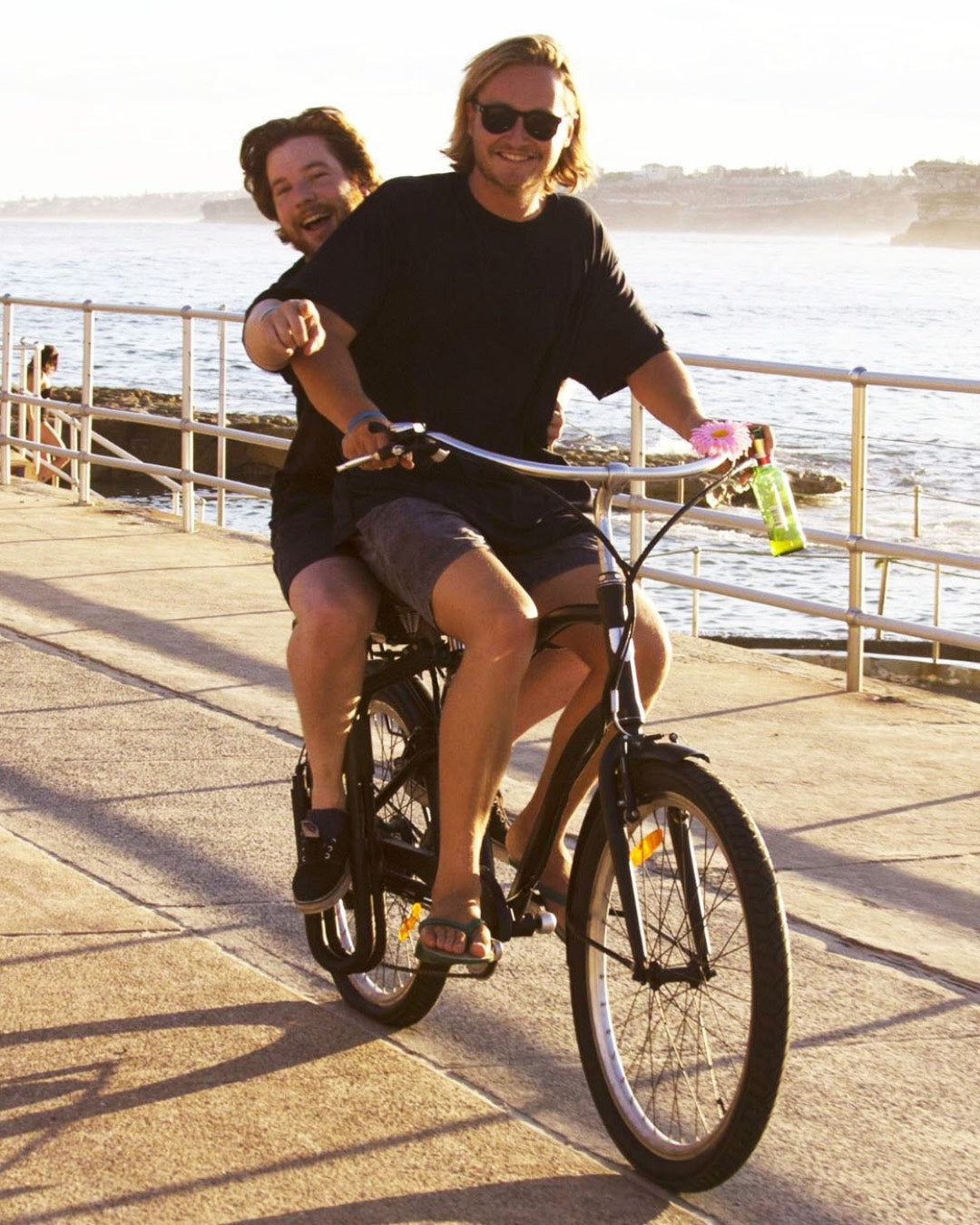 Michael and Conrad, founder of Vamos Bikes Australia enjoying a ride together on a black Vamos Bikes electric bicycle along a sunny coastal boardwalk