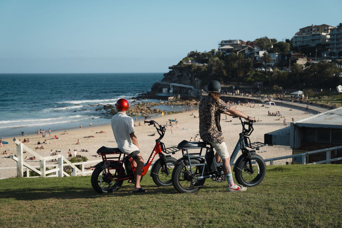 Two people riding Vamos Bikes electric bikes near a scenic coastal beach.