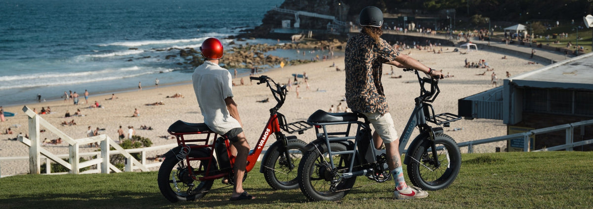 Two people riding Vamos Bikes electric bikes near a scenic coastal beach.