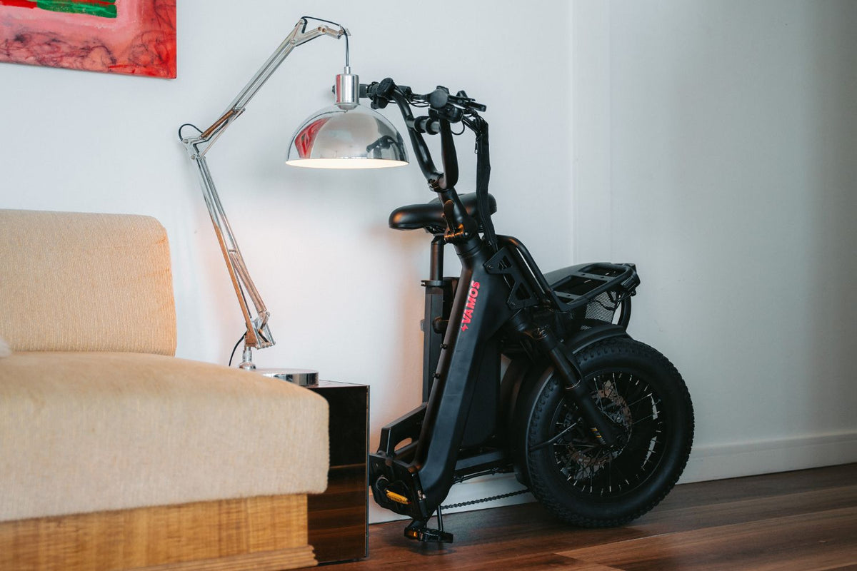 Matte black folded Vamos electric bike stored indoors next to a floor lamp and sofa.