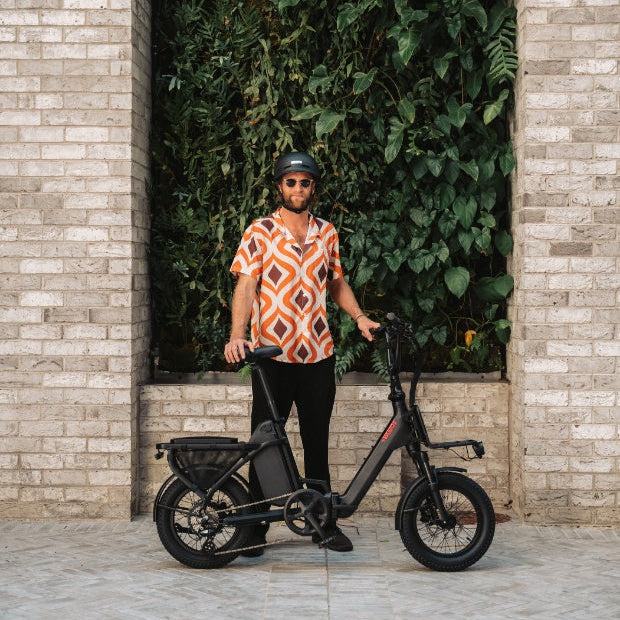 A man in a patterned shirt and helmet stands next to a black Vamos Doblez folding electric bike