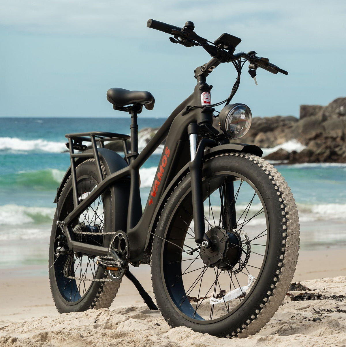 The El Hefe Dual Motor Cruiser Ebike standing on a sandy beach with ocean waves in the background