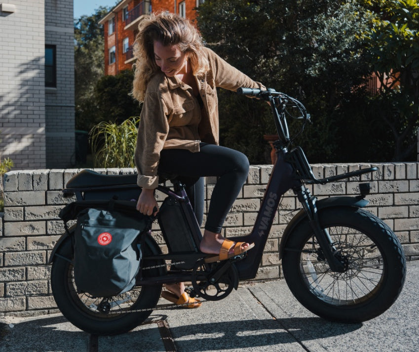 A woman steps onto a Vamos compact cargo electric bike equipped with a Vamos Waterproof Pannier Bag for Bike attached to the rear rack
