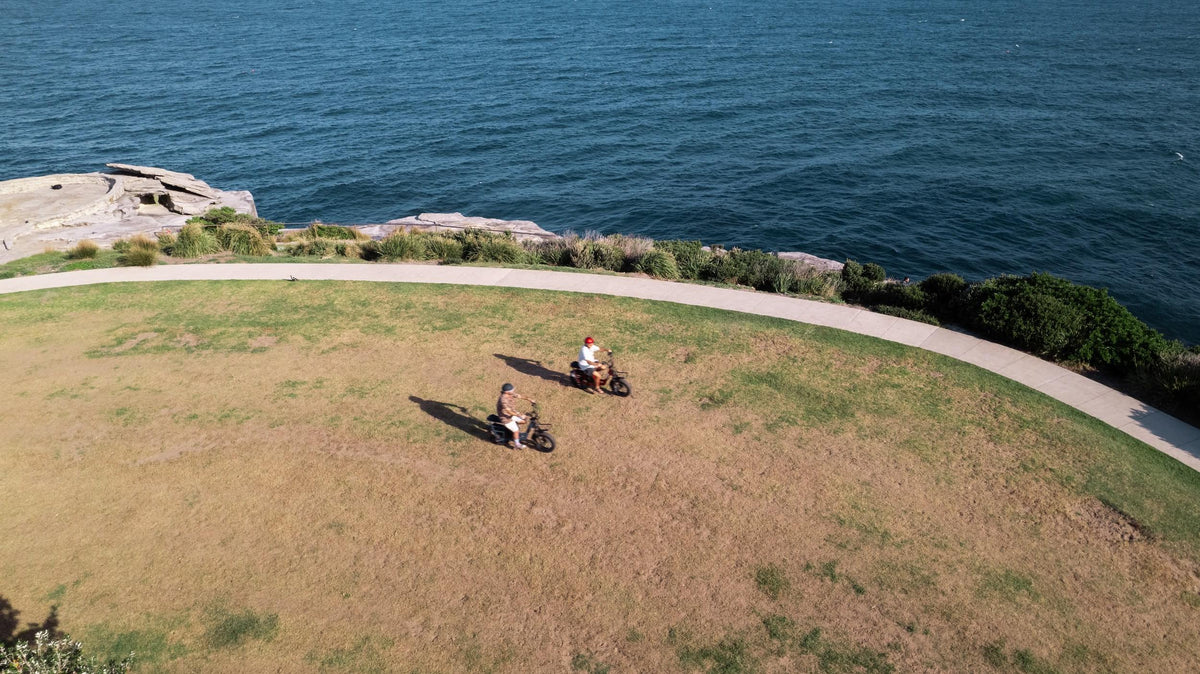 An aerial view of two people riding Vamos Amigo electric bikes on a grassy park overlooking the ocean