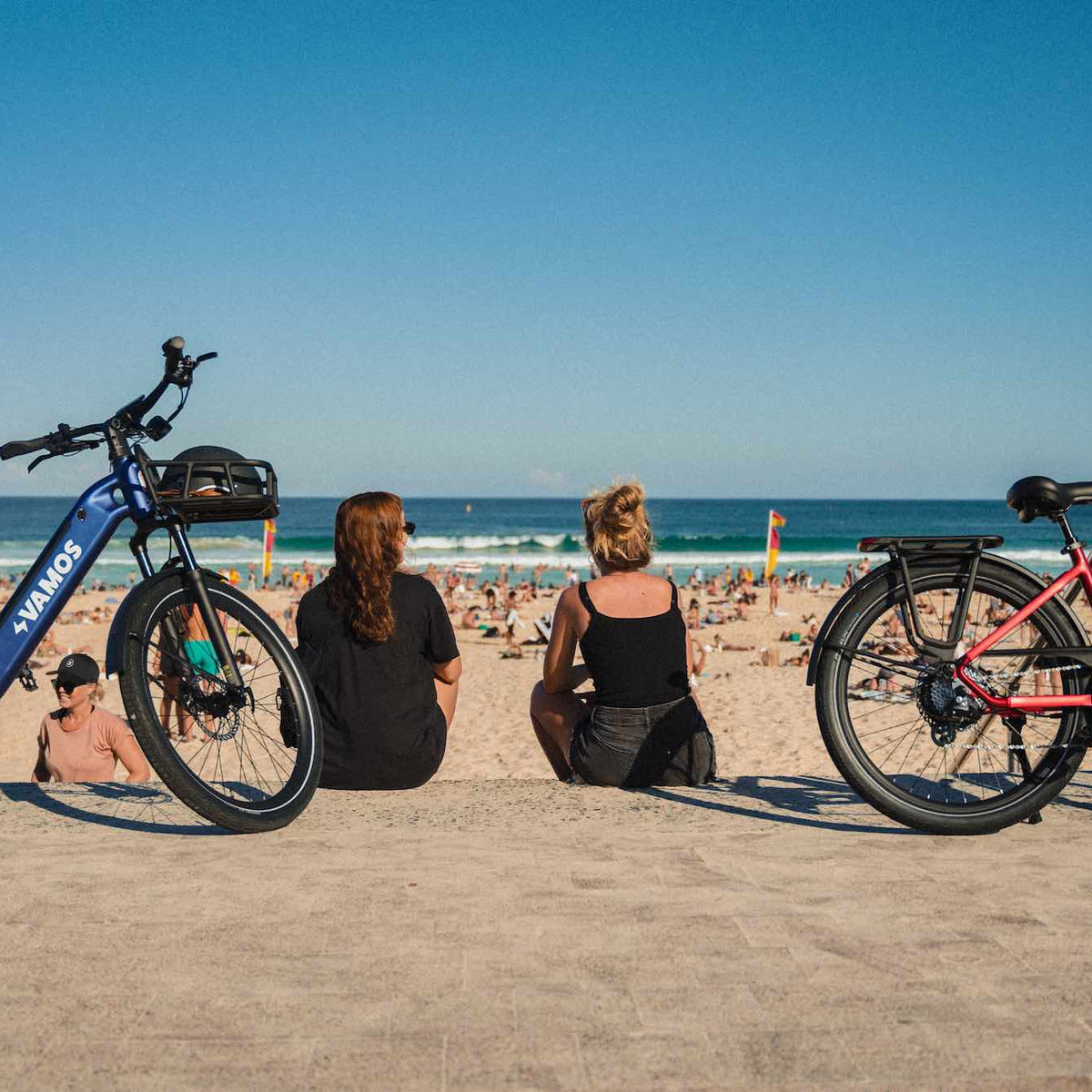 Two Vamos electric bikes parked next to people sitting on a wall overlooking a busy beach.