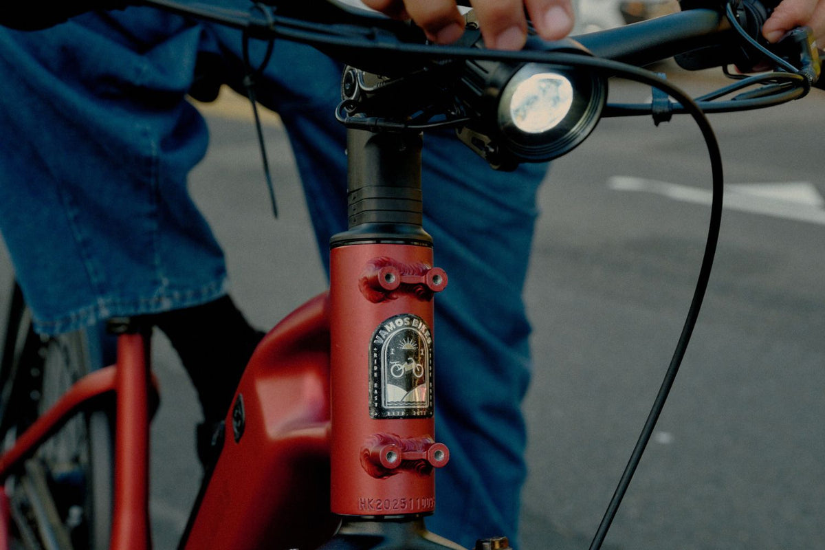 Detail shot of a red Vamos e-bike head tube showing the brand emblem, rack mounts, and handlebar-mounted LED headlight.