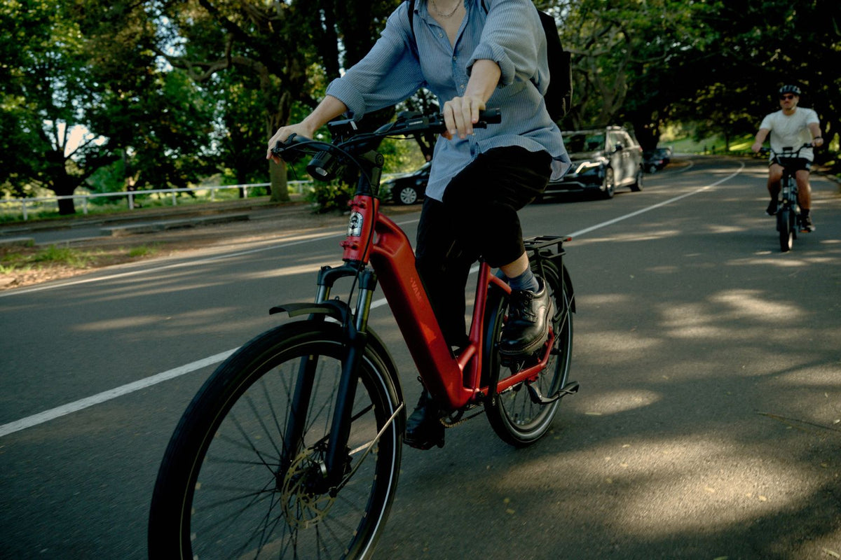 Front-three-quarter angle of a rider on a red step-through Vamos electric commuter bike riding on an urban park road.