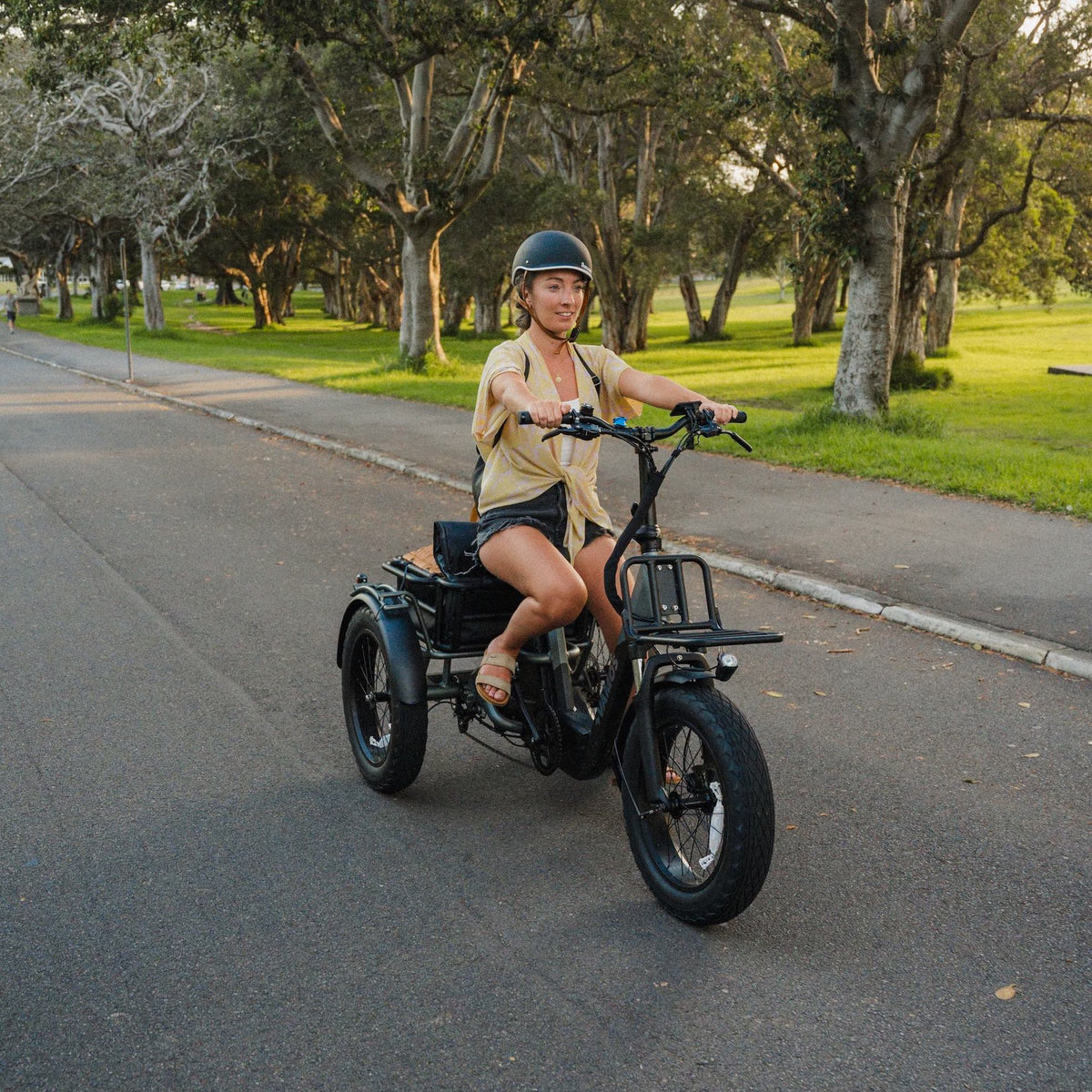 A woman riding a black Vamos Bikes Papa Grande Pro 3-wheel electric trike along a scenic, tree-lined paved path.