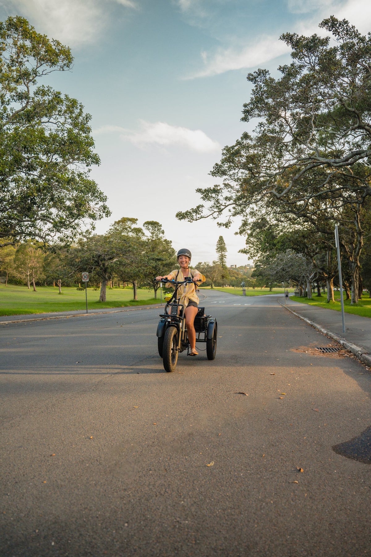A person riding a Vamos Bikes Papa Grande Pro 3-wheel electric trike on a paved roadway lined with green trees.