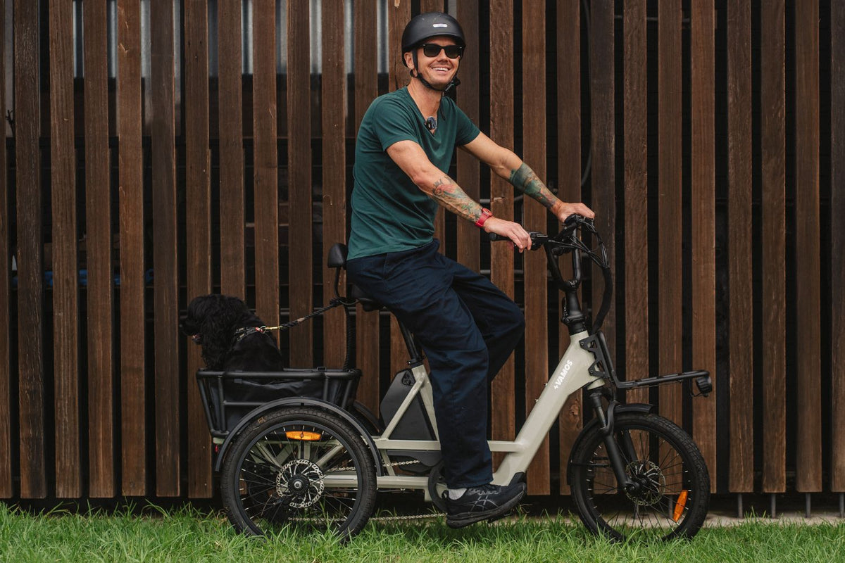 Man posing on a cream-colored Vamos electric cargo tricycle carrying a dog in the rear basket against a wooden fence background.