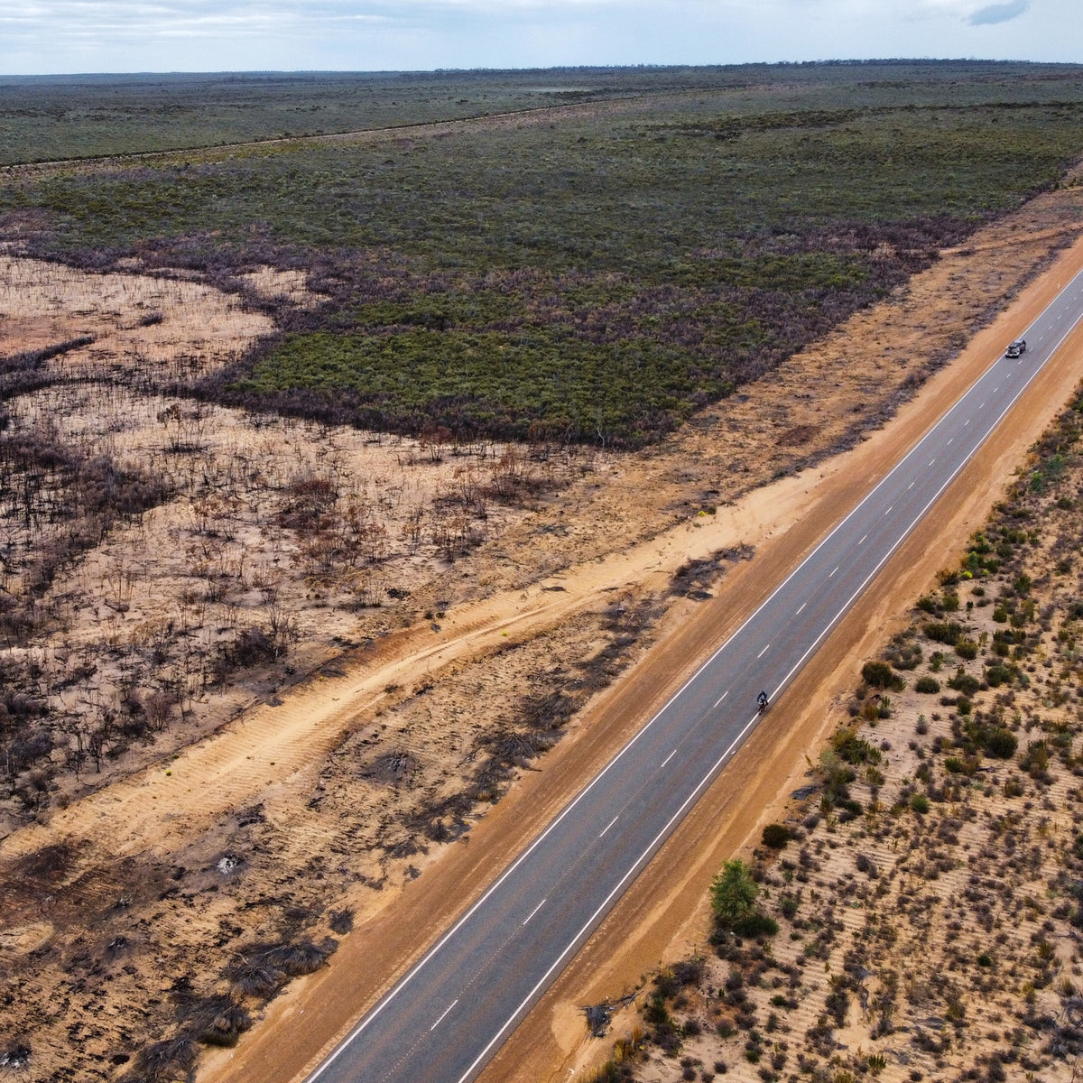 Aerial drone shot of a long, straight paved highway cutting diagonally through a vast, remote landscape with dry shrubland and patches of burnt terrain.