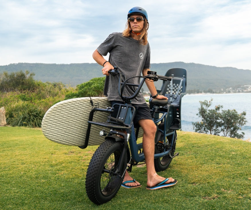 A man stands with an electric cargo bike featuring a surfboard rack and a Thule Yepp 2 Maxi Child Bike Seat mounted on the back.