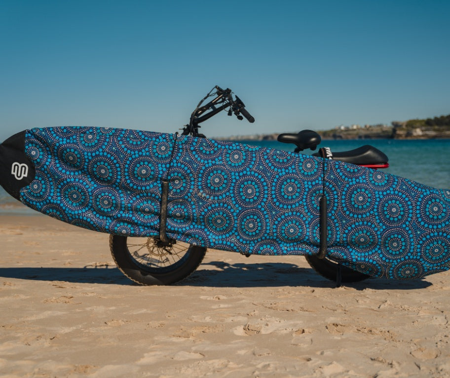 A surfboard in a blue patterned cover mounted on a bicycle parked on a sandy beach.