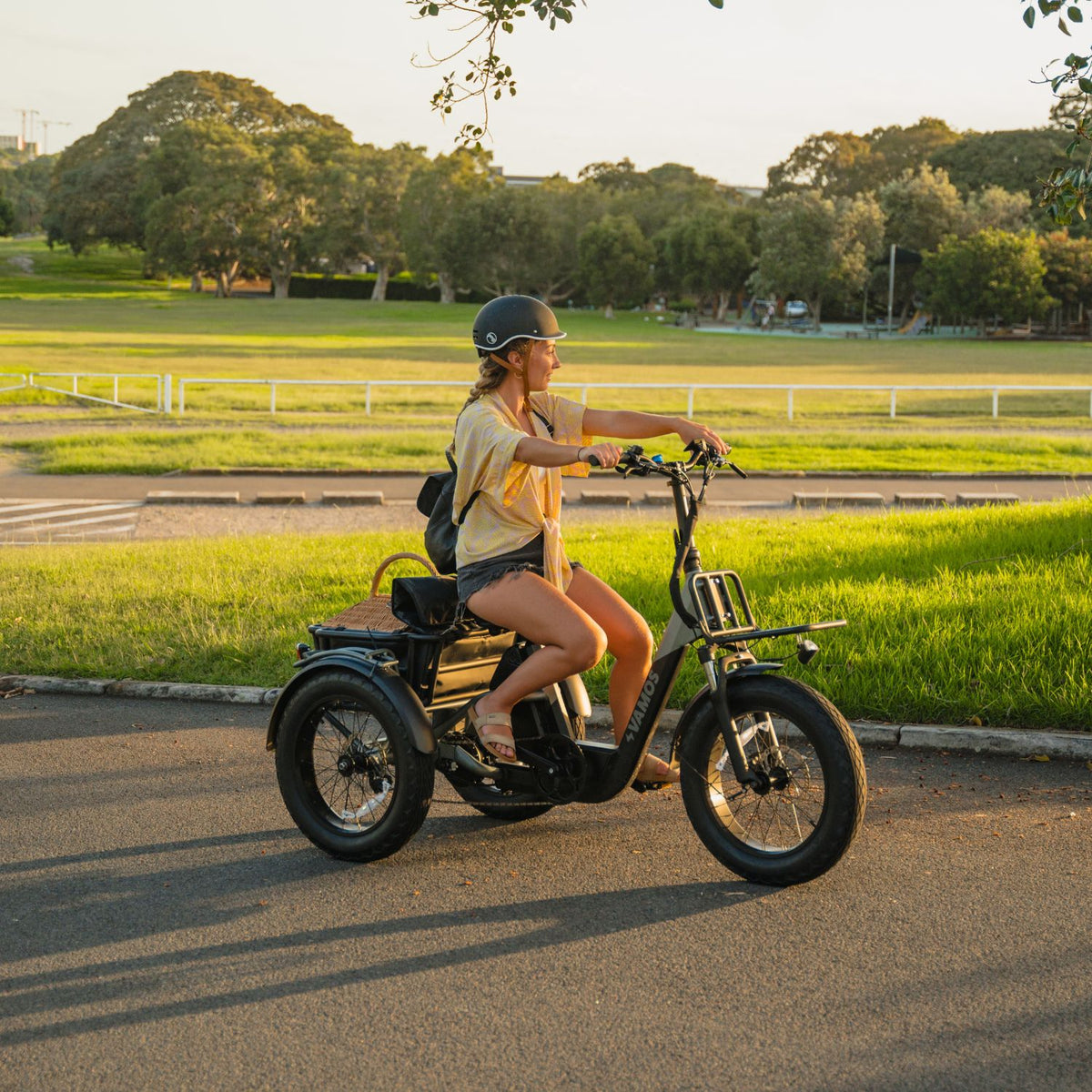 Side view of a woman riding a black Vamos fat-tire electric tricycle with front rack and rear cargo basket in a park.