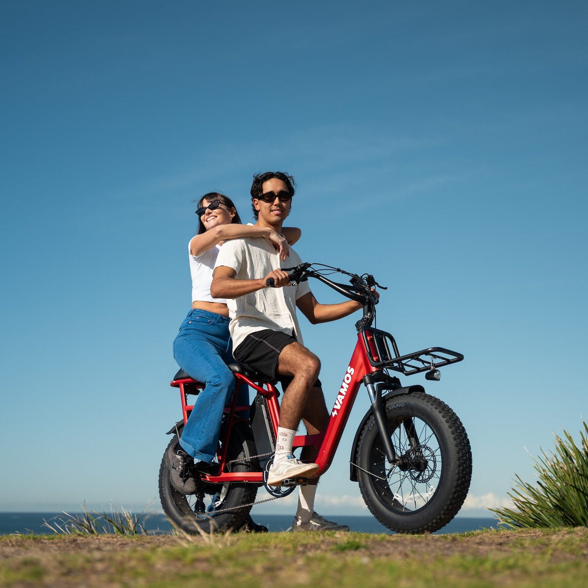 A couple riding a red Vamos fat-tire electric cargo bike on a grassy hill overlooking the ocean against a clear blue sky.