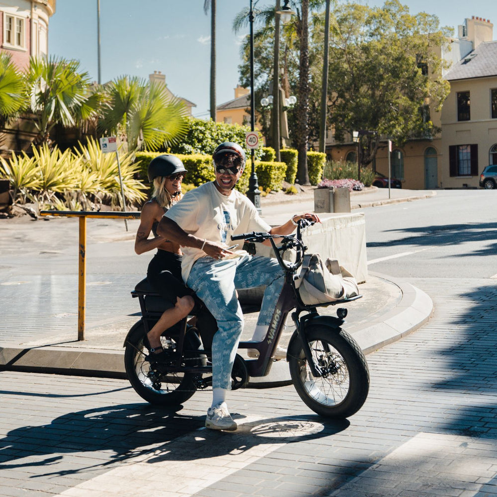 A person riding a Vamos El Amigo compact cargo e-bike with fat tires while a passenger sits on the back.