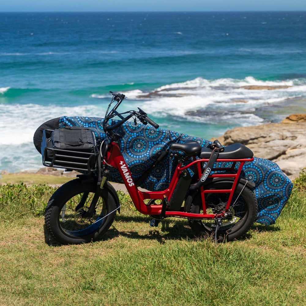 Red Vamos El Amigo compact cargo e-bike equipped with a front rack and a surfboard rack carrying a surfboard along the coast.