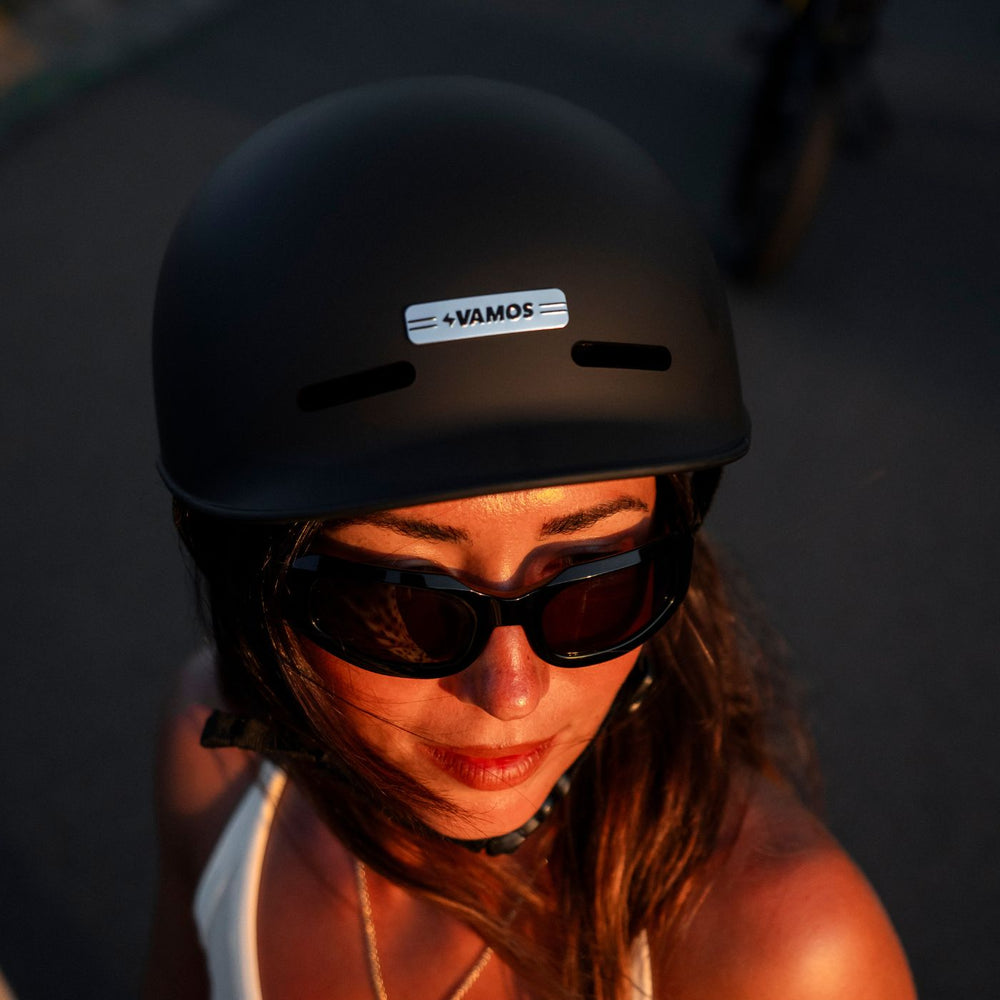 A person wearing a matte black Vamos Bike Helmet featuring a reflective brand logo on the front.