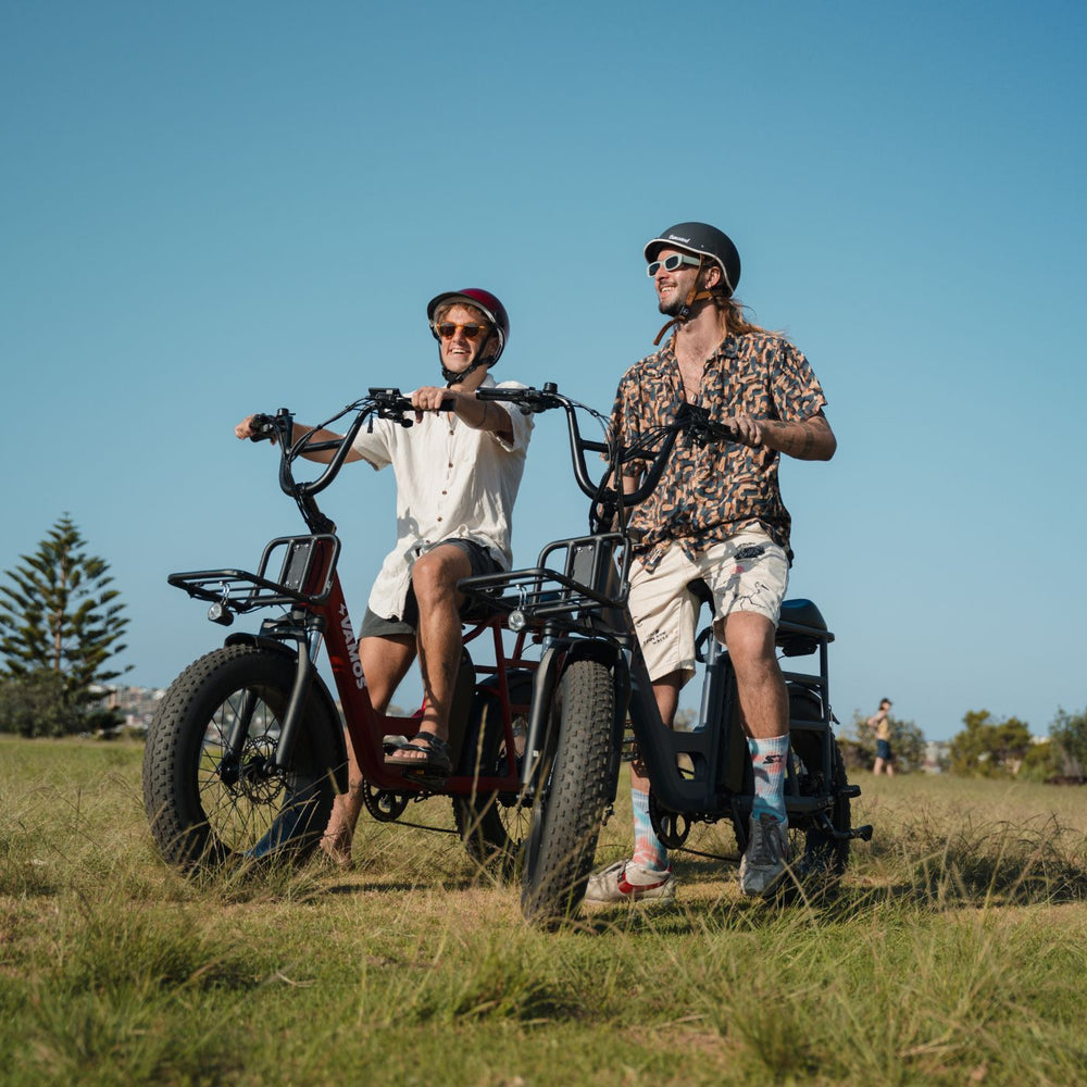 Two people riding Vamos electric cargo bikes with fat tires on a grassy field under a clear blue sky.