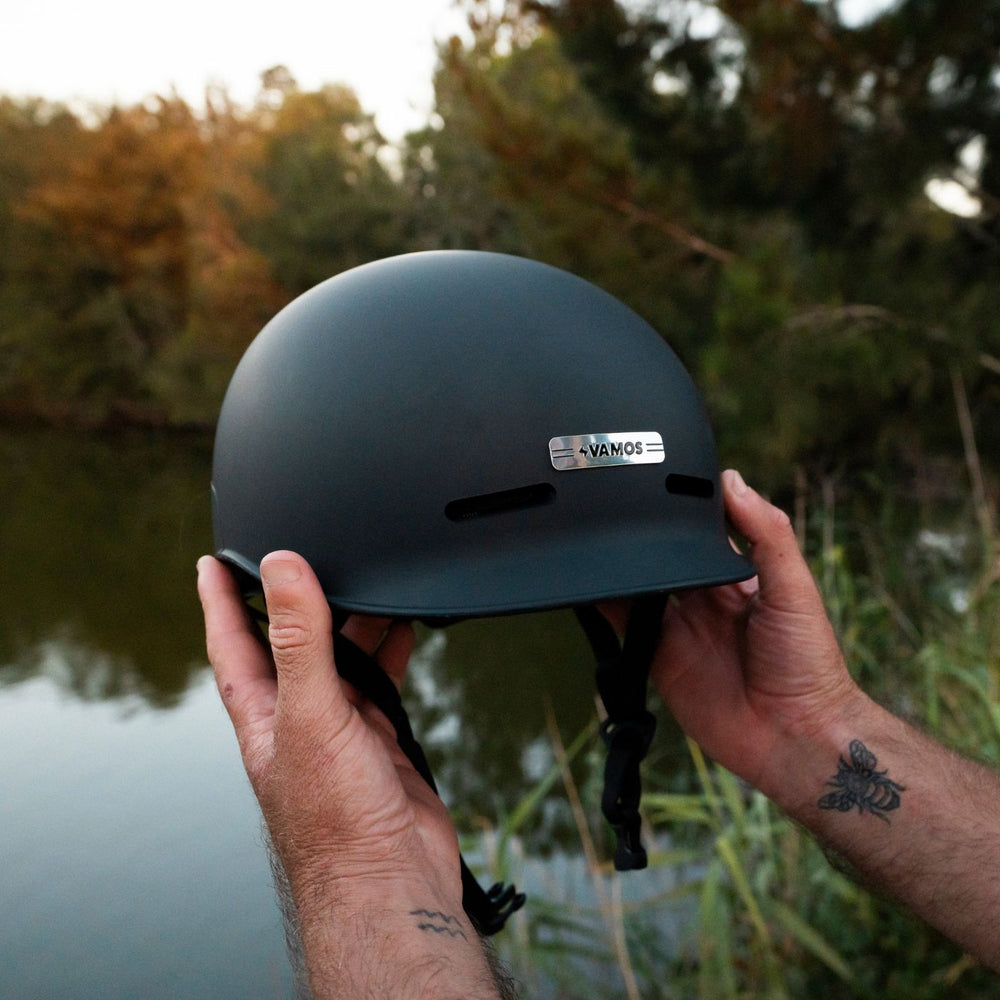A matte black Vamos e-bike helmet featuring a silver-branded metal badge on the front.