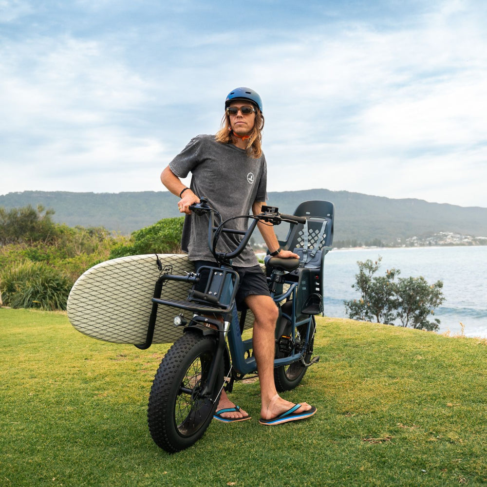 A person posing next to a Vamos electric fat bike equipped with a front-mounted surfboard rack and a rear child seat.