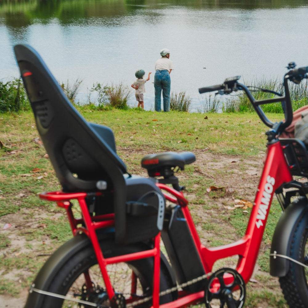 A red Vamos Amigo electric cargo bike equipped with a black Thule Yepp 2 Maxi child bike seat parked near a grassy lake shore.
