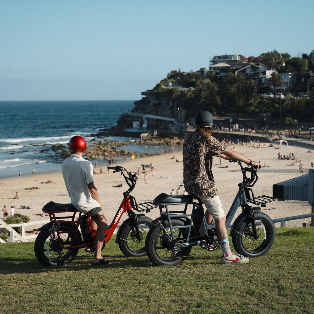 Two people riding Vamos Bikes electric bikes near a scenic coastal beach.