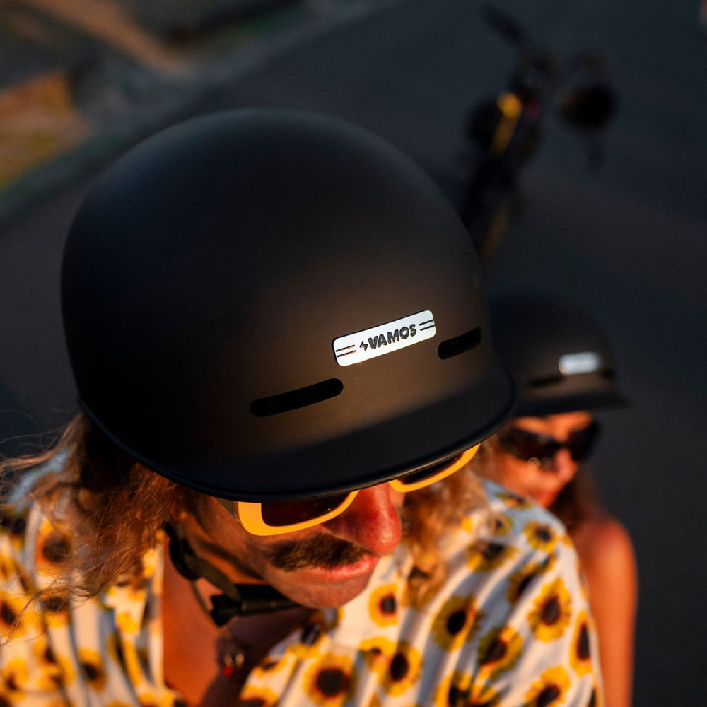 A person wearing a matte black Vamos Bike Helmet featuring a reflective "VAMOS" badge on the front.