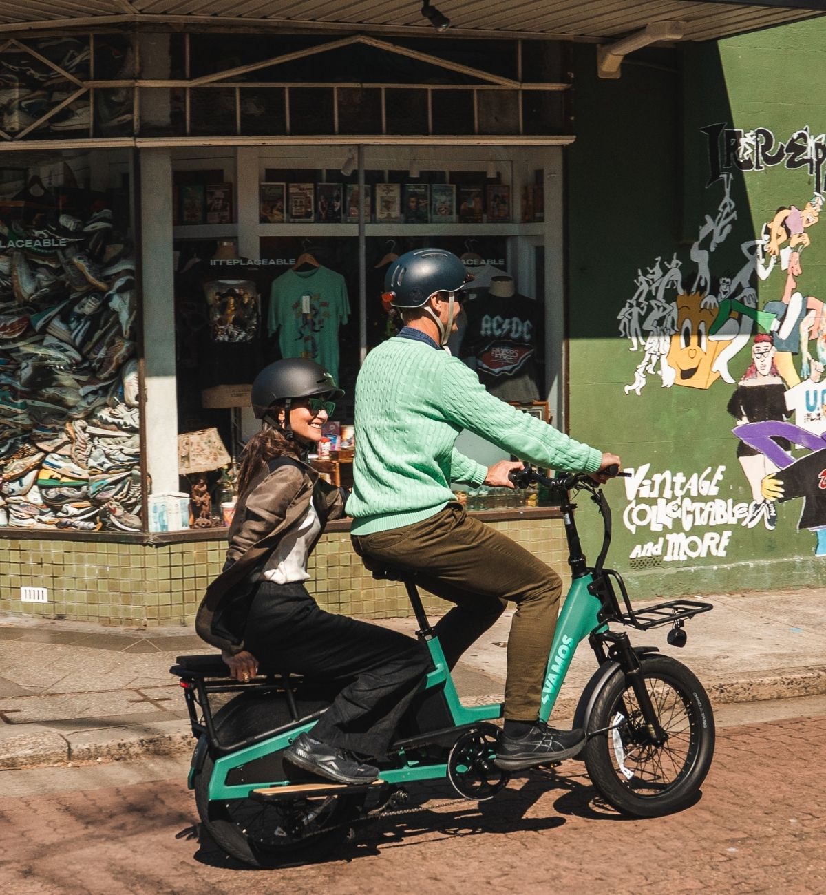 A man and woman wearing helmets ride a Vamos Amigo Plus green electric cargo and passenger bike. past a vintage shop