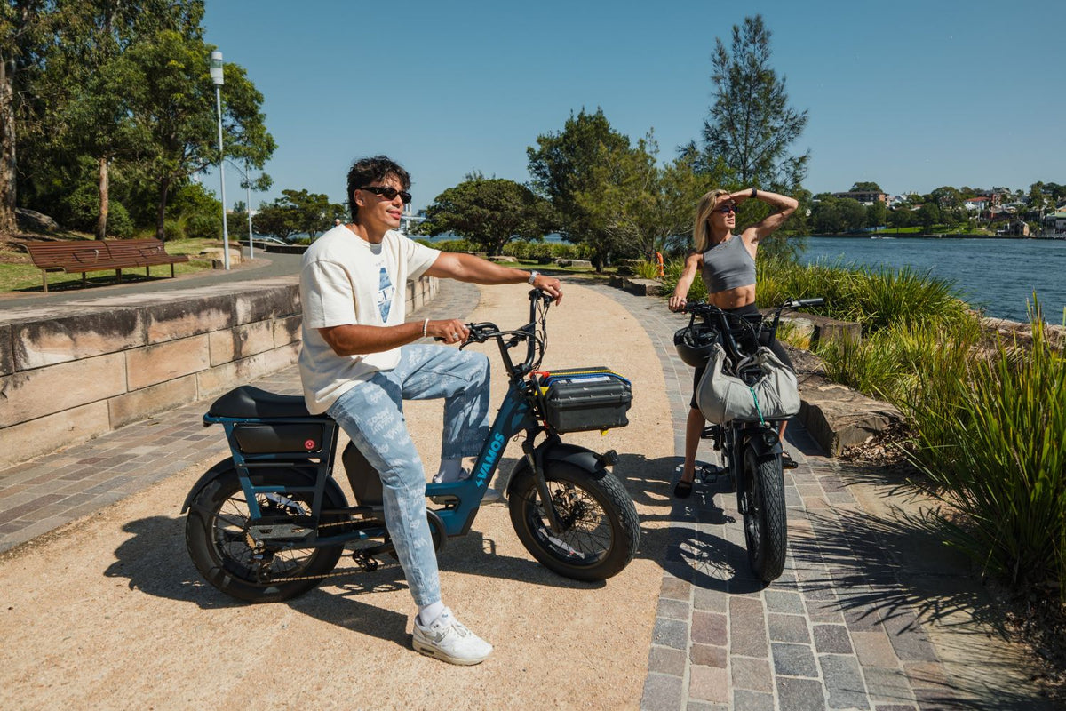 A person rides a Vamos Amigo Plus 2026 All-Purpose Passenger Commuter E-Bike along a paved waterfront path next to a passenger on a sunny day.