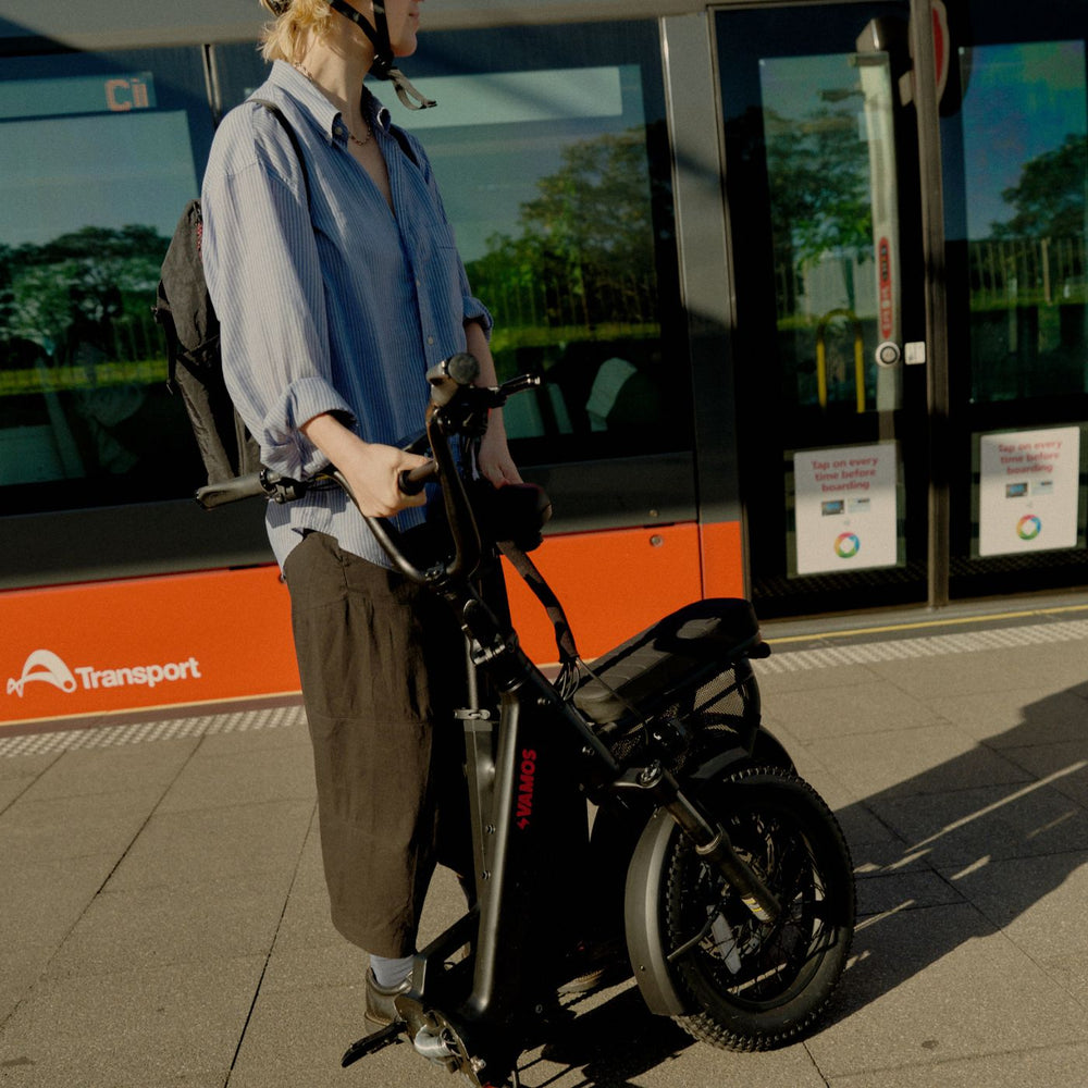 Commuter standing with a folded black Vamos electric bike on a public transport platform.