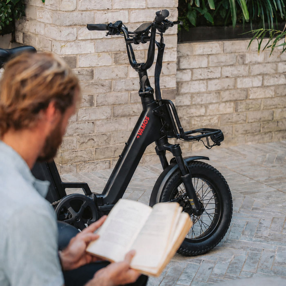 Man reading a book outdoors next to a matte black Vamos compact electric bike with a front rack.