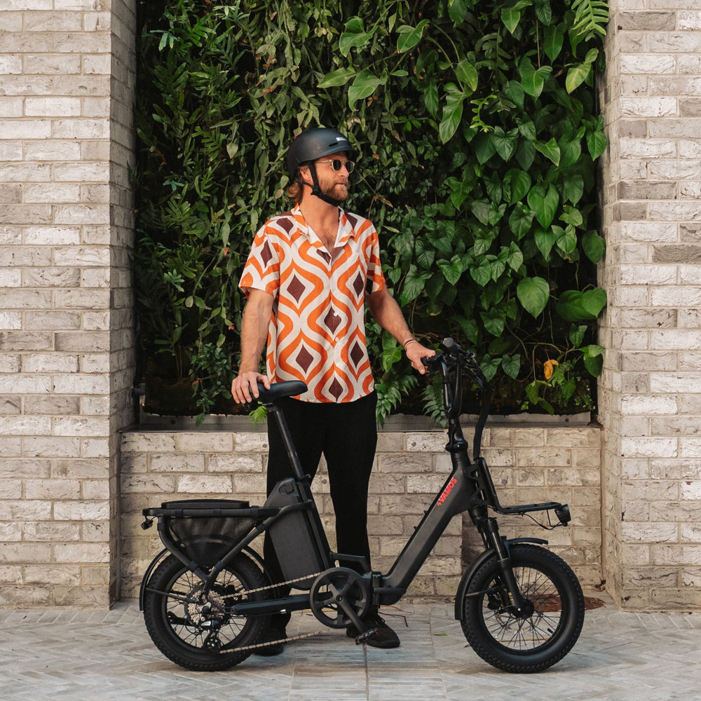 Man in a patterned shirt and helmet standing with a matte black Vamos compact electric bike in front of a green wall feature.