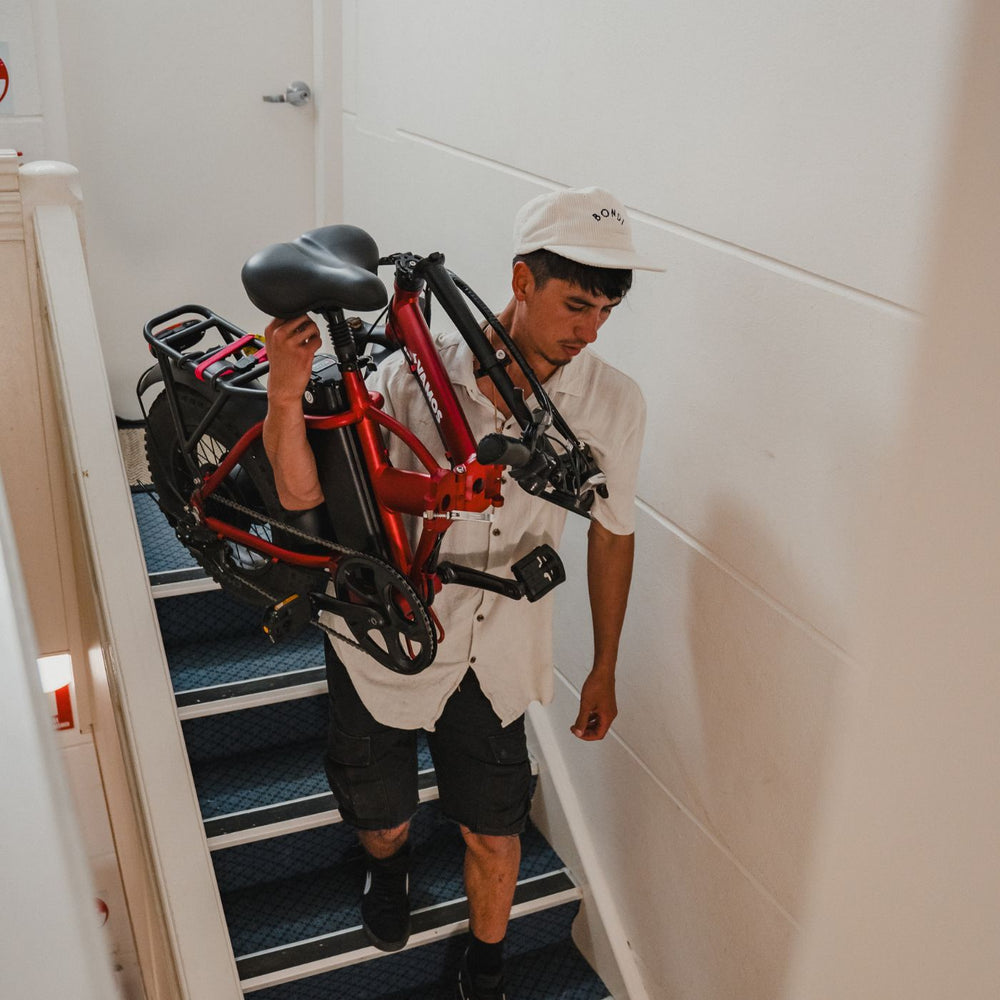 Man carrying a folded metallic red Vamos electric bike up a set of stairs inside a building.