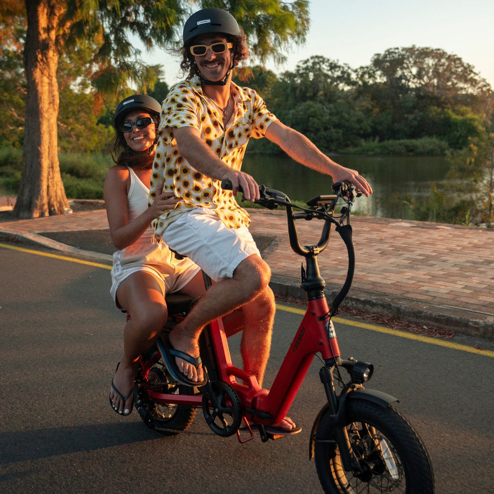 Couple wearing helmets riding together on a red double-seat Vamos electric bike along a paved park path.
