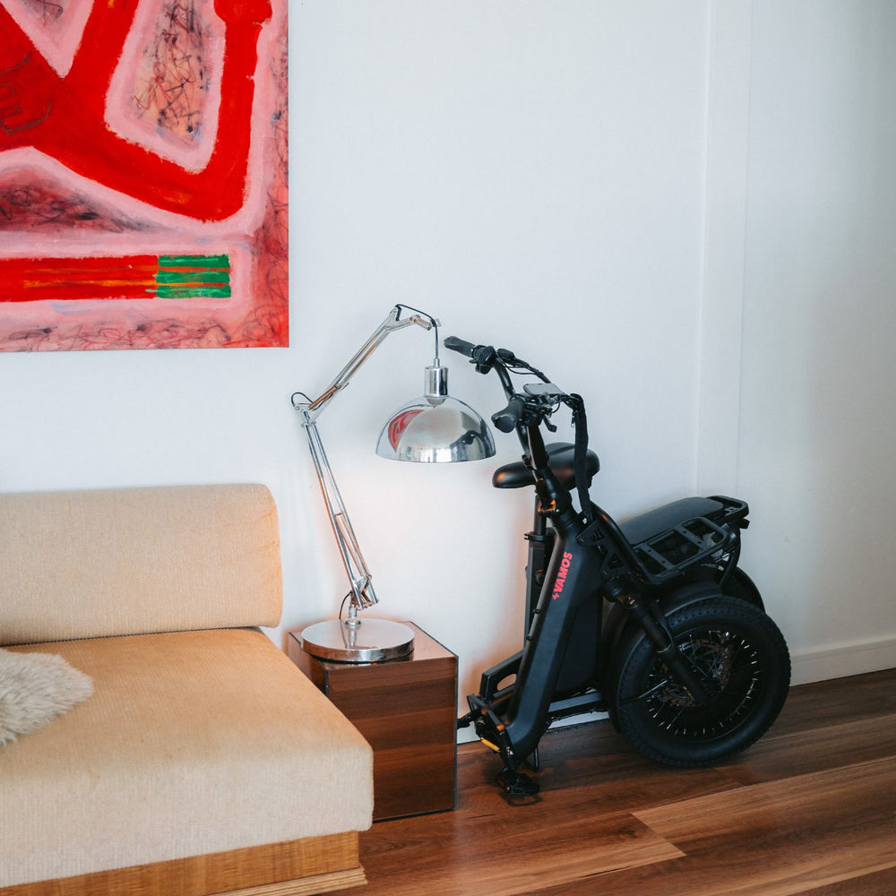 Matte black folded Vamos electric bike standing indoors next to a side table and sofa with artwork in the background.