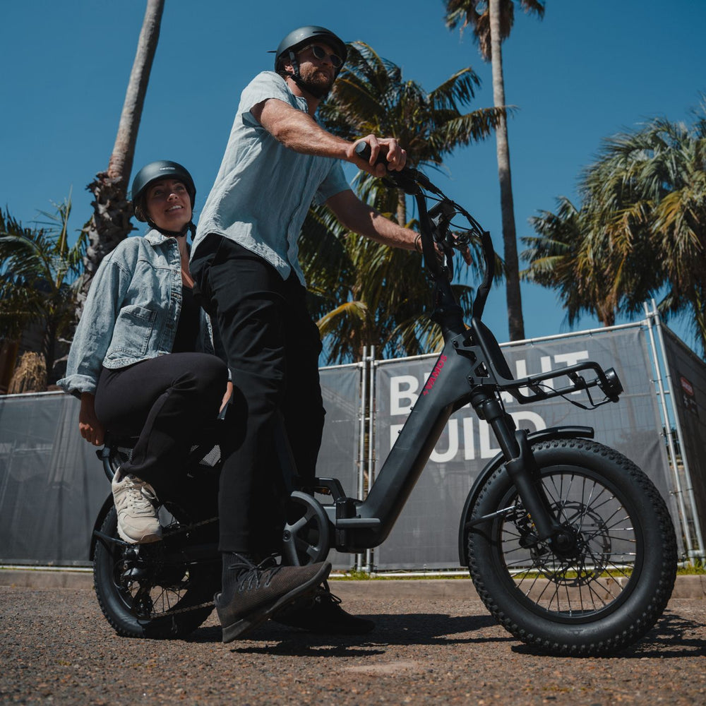 Couple riding together on a black double-seat Vamos electric bike under palm trees.