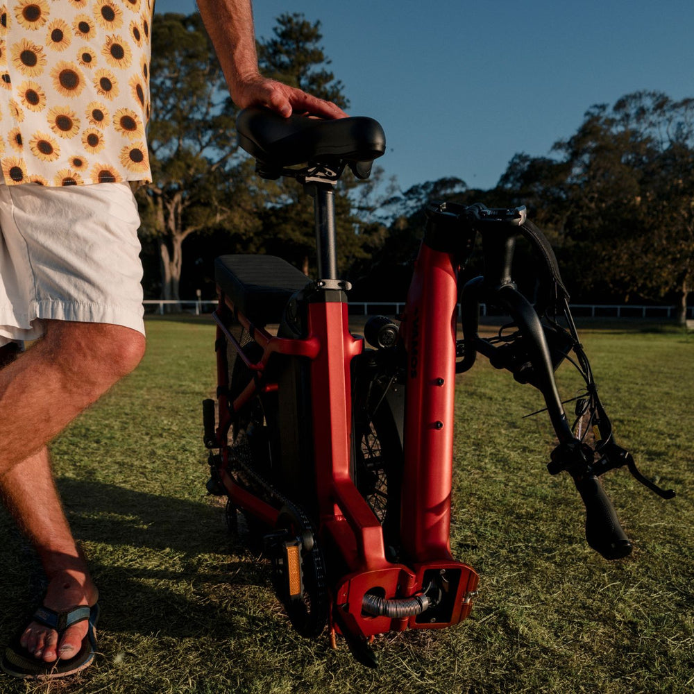 Man standing beside a folded metallic red Vamos electric bike on a grassy field.
