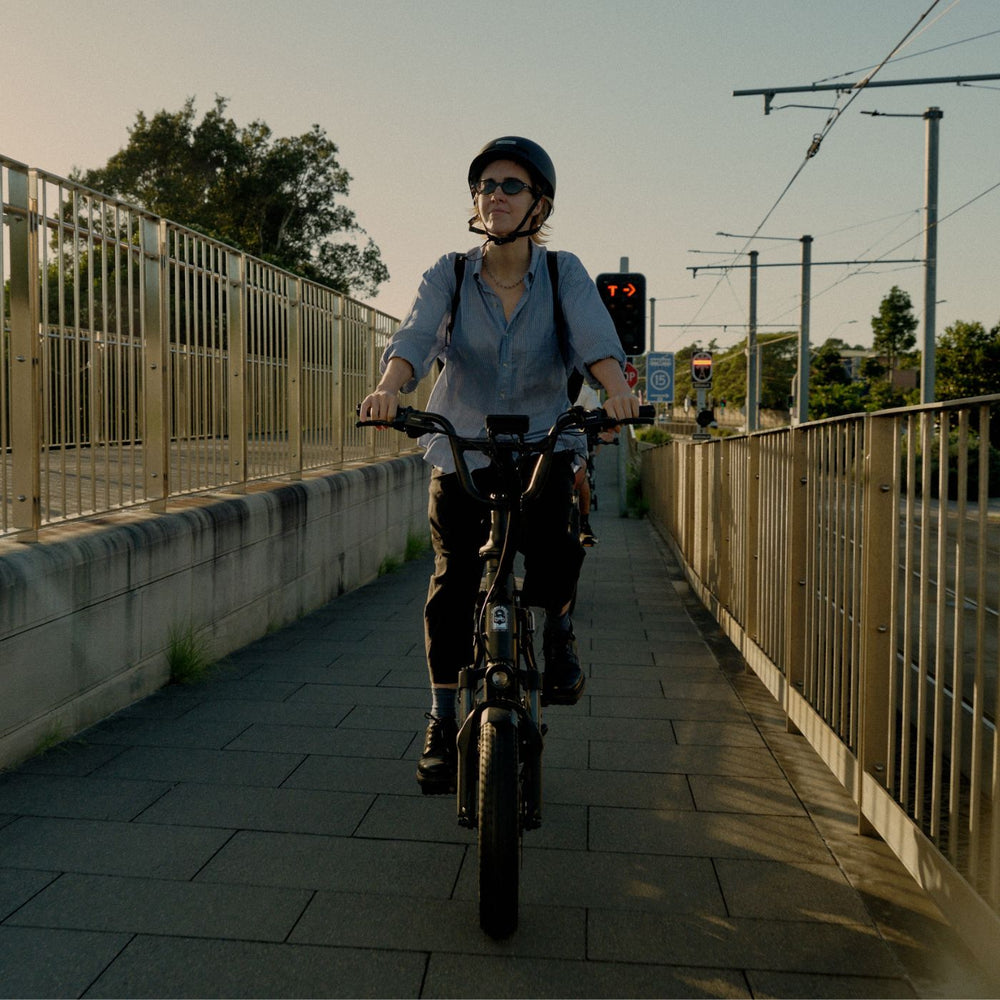 Woman wearing a helmet riding a black Vamos electric bike along a fenced urban pathway.