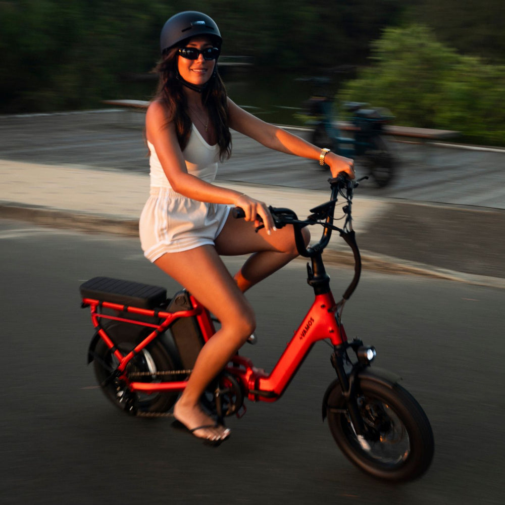 Woman wearing a helmet riding a bright red compact Vamos electric bike along a paved road at sunset.