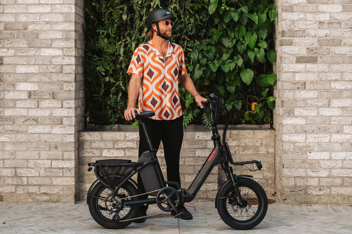 Man wearing a helmet standing next to a black Vamos electric bike against a brick wall feature with greenery.