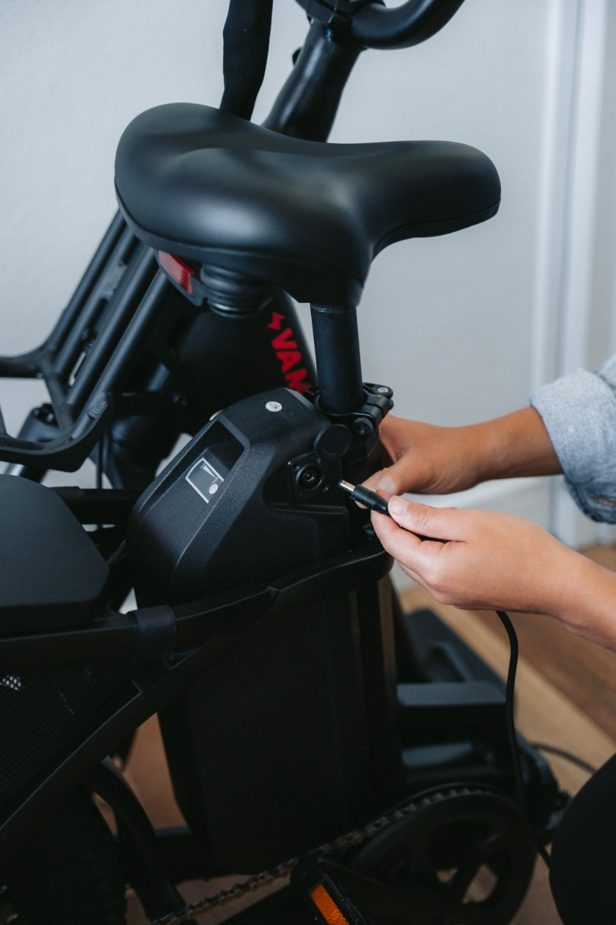 A person plugging a charger cable into the battery pack mounted below the seat of an electric bicycle.