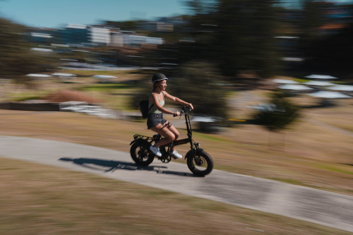 A person riding a compact, black Vamos Doblez 2026 Foldable E-Bike on a paved path.