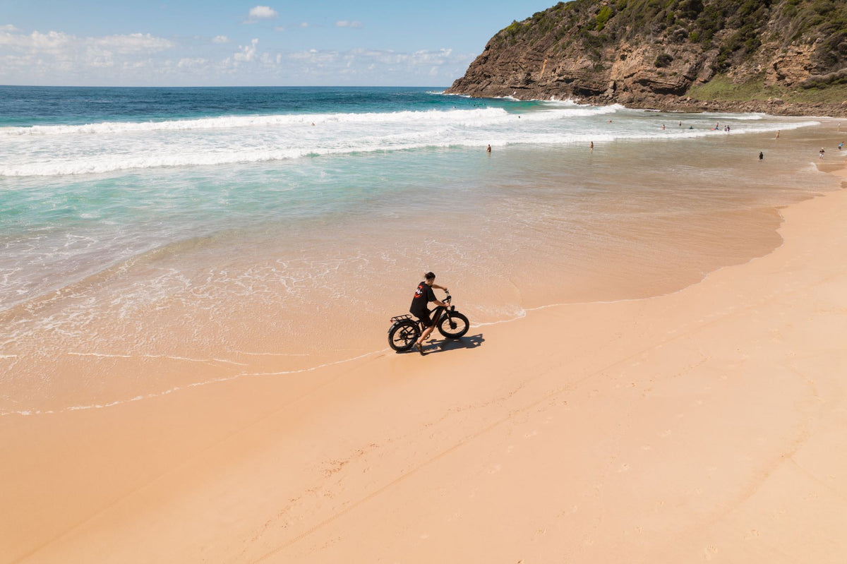 An aerial view of a person riding an El Hefe Dual Motor Cruiser Ebike along the tide line of a beach 