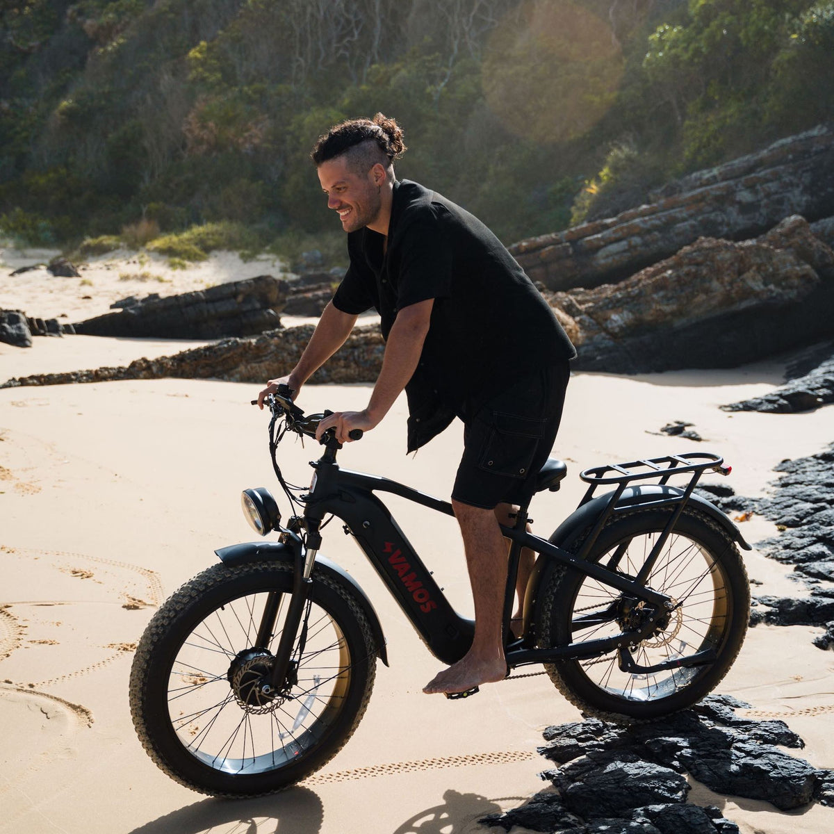 Side view of a man riding an El Hefe Dual Motor Cruiser Ebike over a sandy and rocky beach terrain