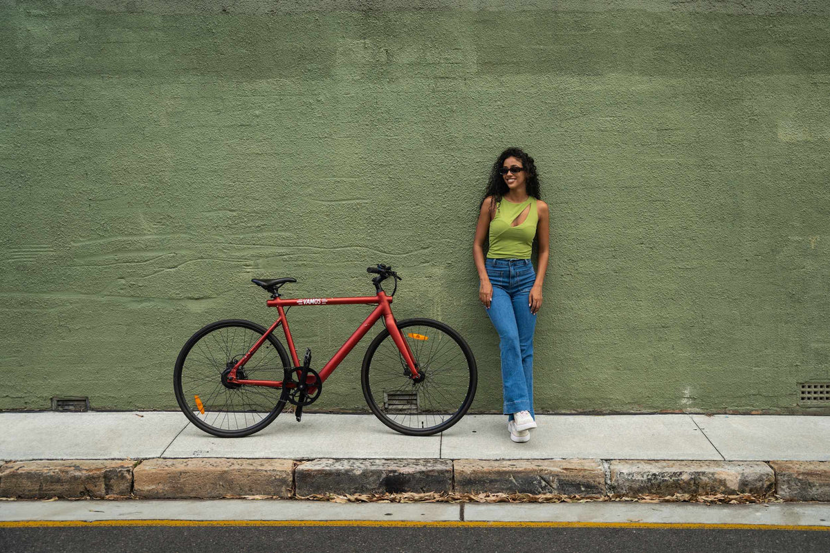 A person standing next to a red Vamos electric bicycle in front of a textured green wall.