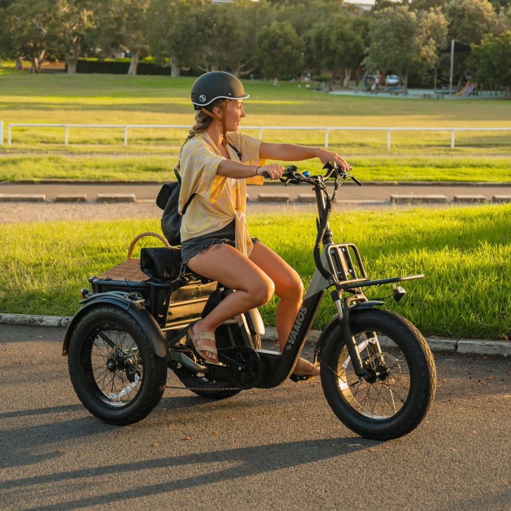 Side view of a woman in a helmet riding a black Vamos electric trike with a picnic basket at golden hour.