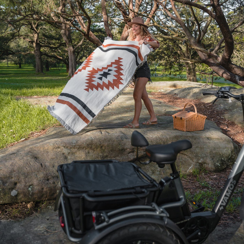 Woman laying out a picnic blanket in a park next to a black Vamos electric tricycle with rear cargo basket.