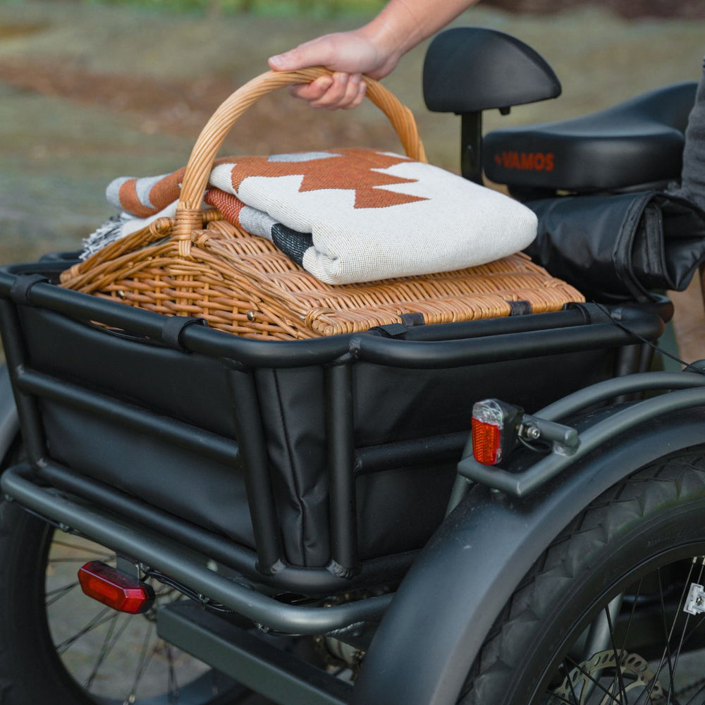 Hand placing a wicker picnic basket and folded blanket into the rear cargo rack of a black Vamos electric tricycle.