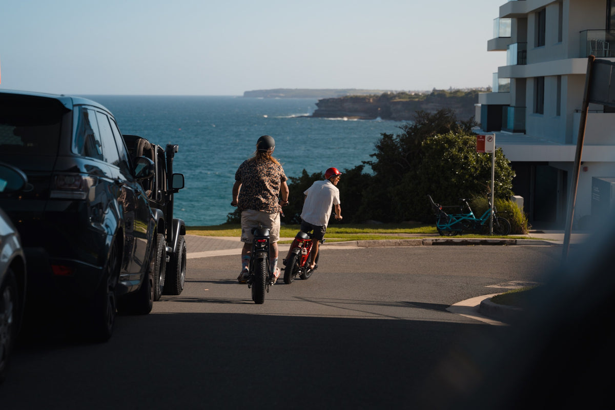 Two riders on Vamos electric bikes riding downhill toward the ocean in a coastal Australian suburb